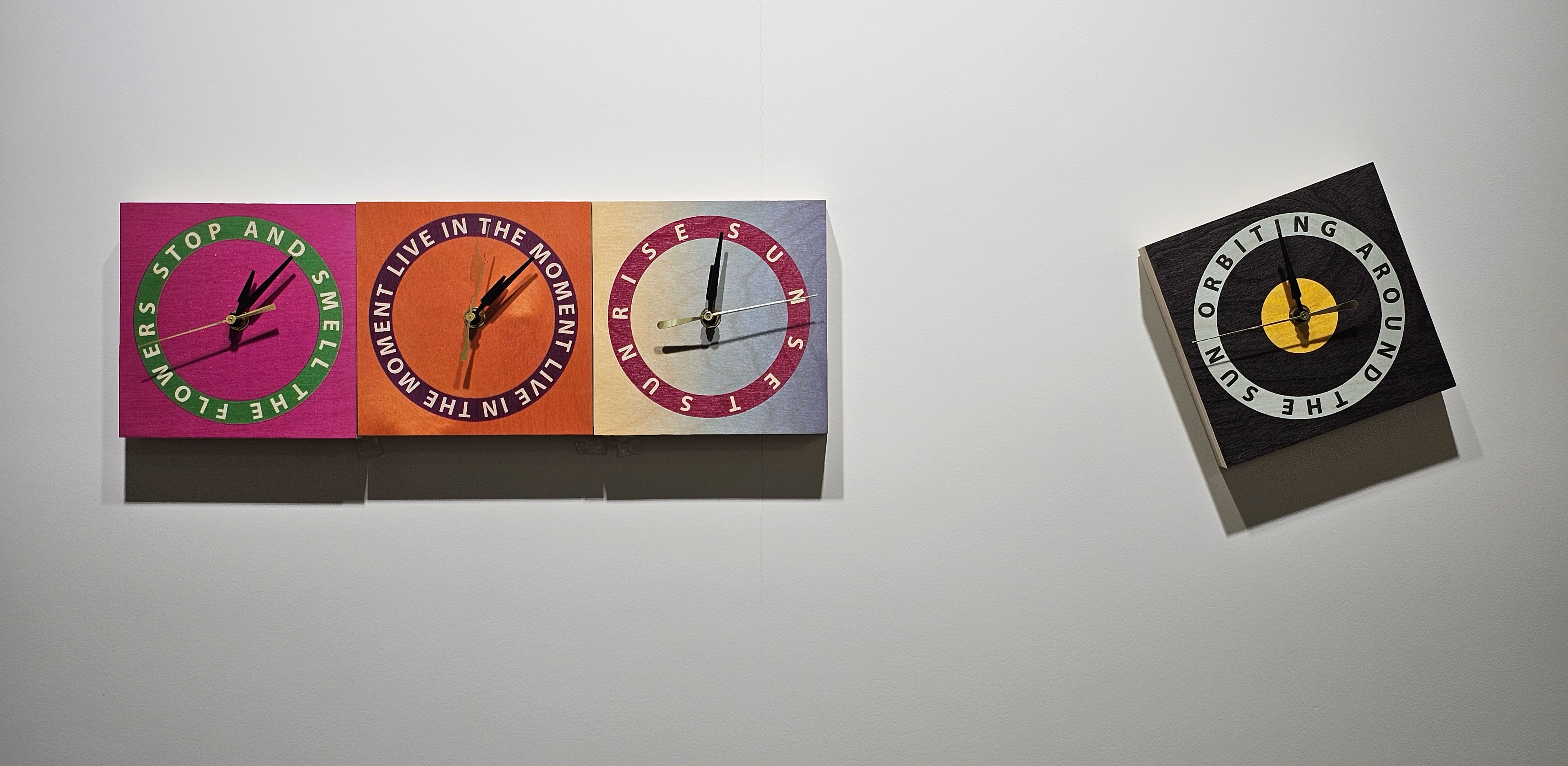 A wall with two artworks: one rectangular piece with four colorful clock faces and another smaller square piece with a single clock face.
