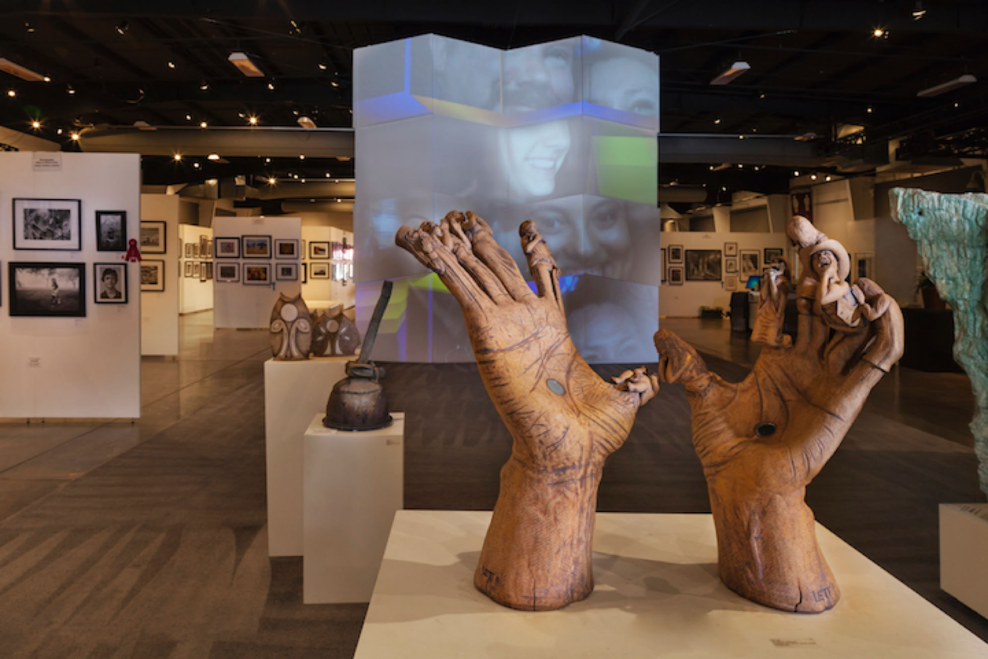 Sculpture of two large hands displayed in an art gallery with various artworks on the walls and a large screen in the background.