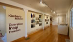 A gallery room with wooden floors and white walls displaying photographs. The wall on the left has text reading "Kimberly Millett's Operation Iraqi Freedom." The room is lit with track lighting, and there is a display case on the right.